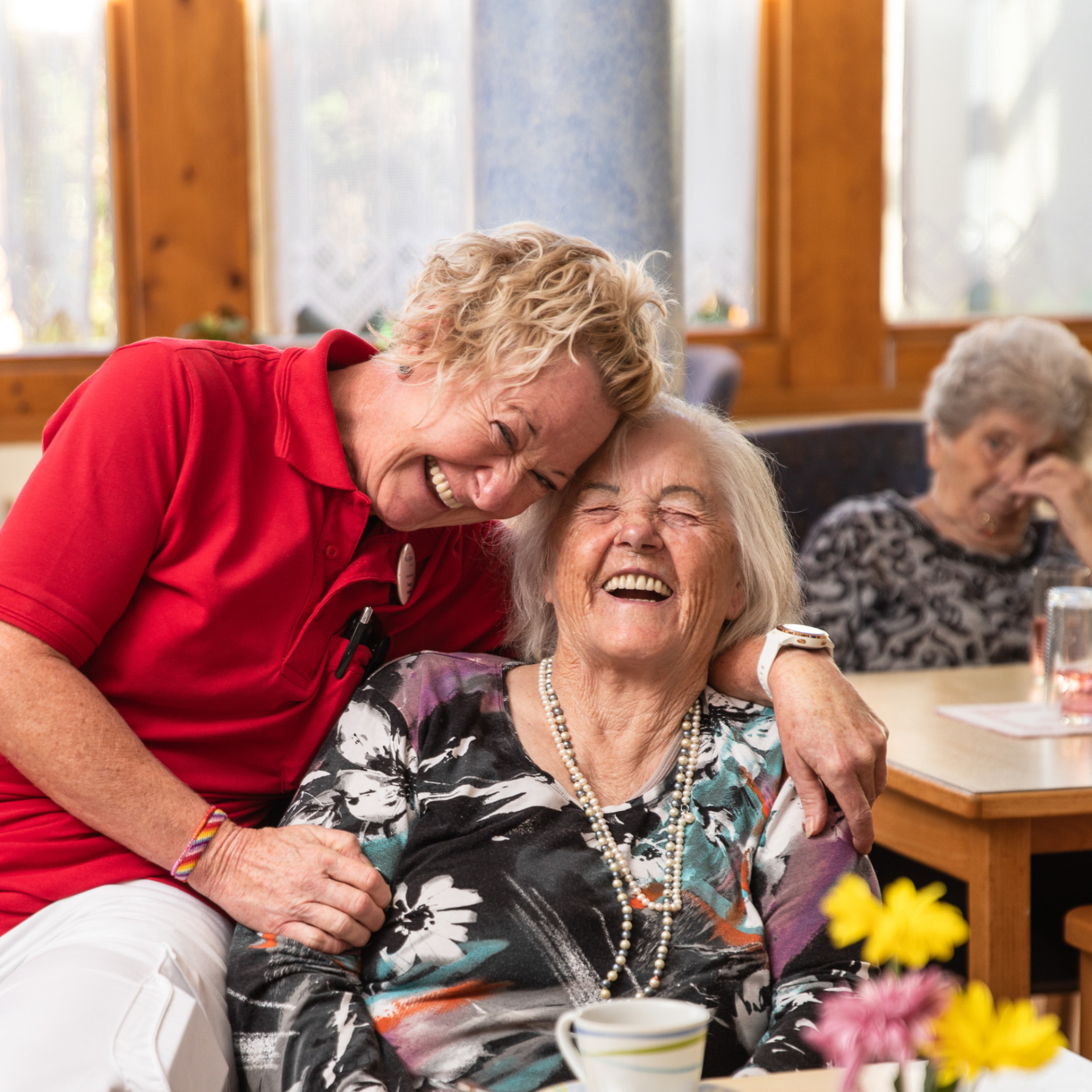 Mitarbeiterin legt liebevoll den Arm um eine lachende Bewohnerin, während beide gemeinsam an einem gedeckten Tisch sitzen und lachen.