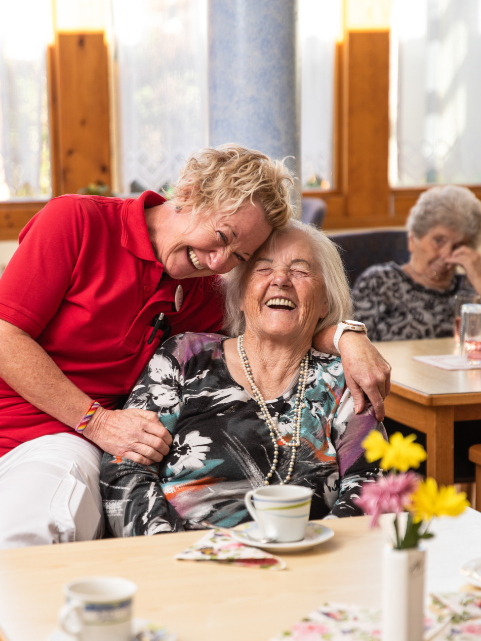 Mitarbeiterin legt liebevoll den Arm um eine lachende Bewohnerin, während beide gemeinsam an einem gedeckten Tisch sitzen und lachen.