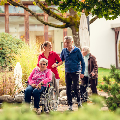 Bewohnerinnen und Bewohner spazieren gemeinsam mit einer Betreuerin durch den Garten, eine Bewohnerin sitzt im Rollstuhl.
